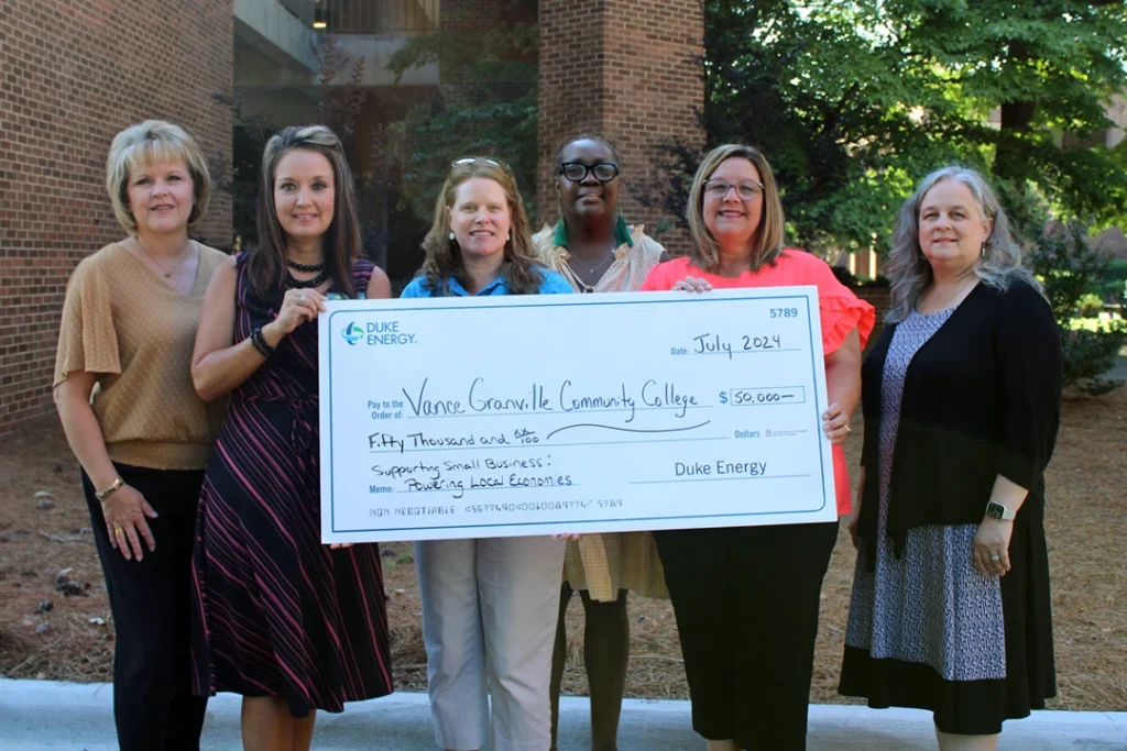 A group of six women posing outdoors with a large presentation check