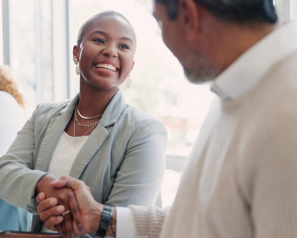 Stock photo; young woman in a blazer shakes hands with a middle-aged man wearing a button-down shirt [Shutterstock ID 2265615103].